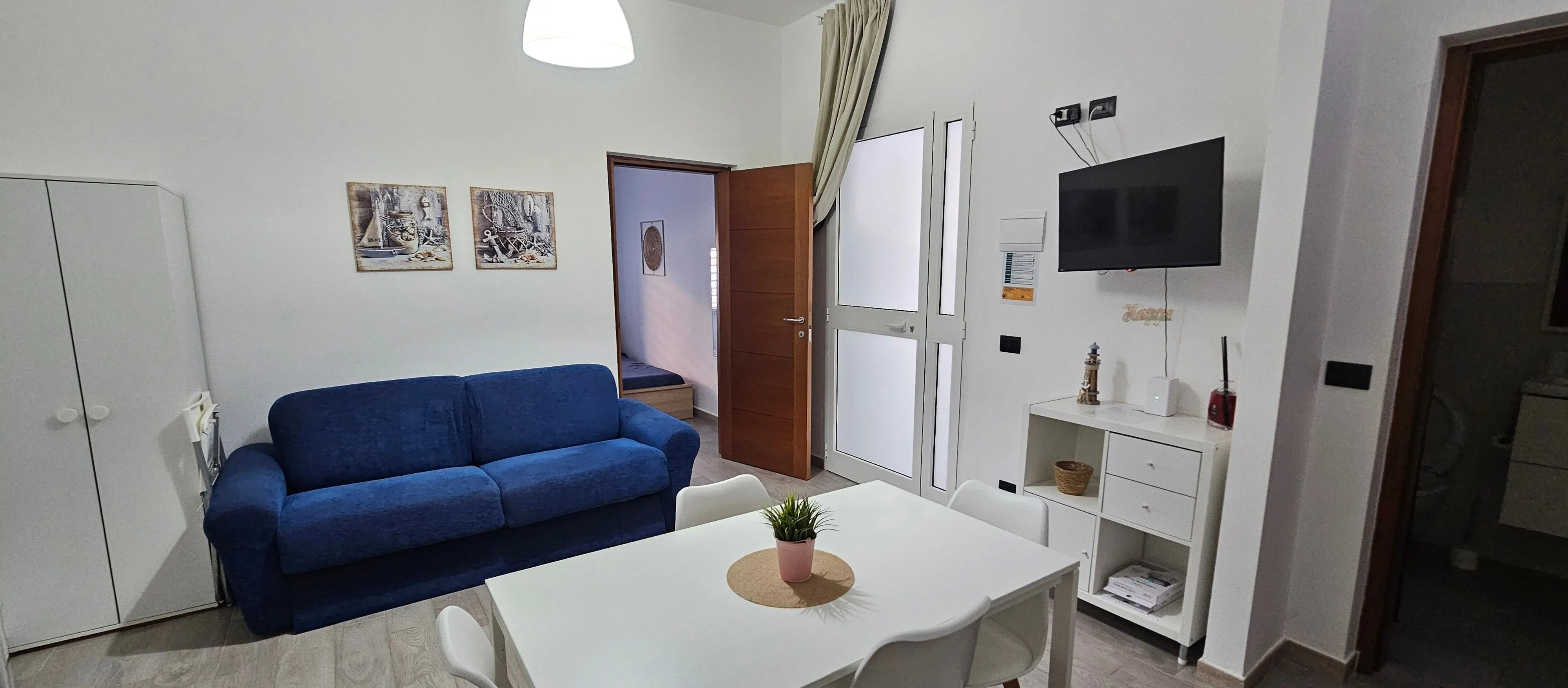 Zona giorno dell'appartamento: divano blu, tavolo bianco, TV e porte interne.